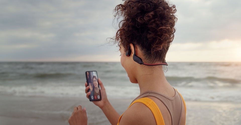 Športovec telefonujúci na pláži prostredníctvom použítia slúchadiel s kostným vedením zvuku